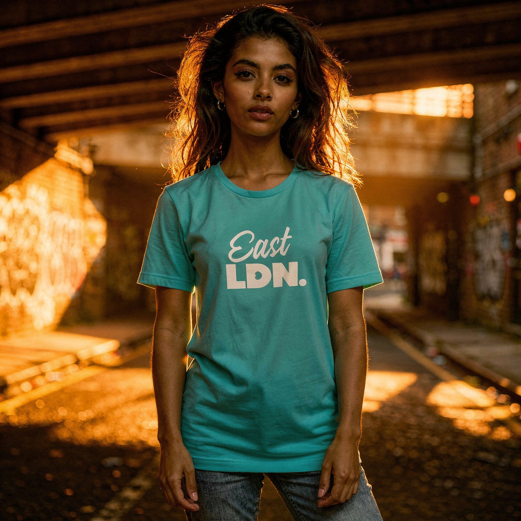 Woman wearing a teal 'East LDN.’ t-shirt standing in an urban setting with warm lighting.