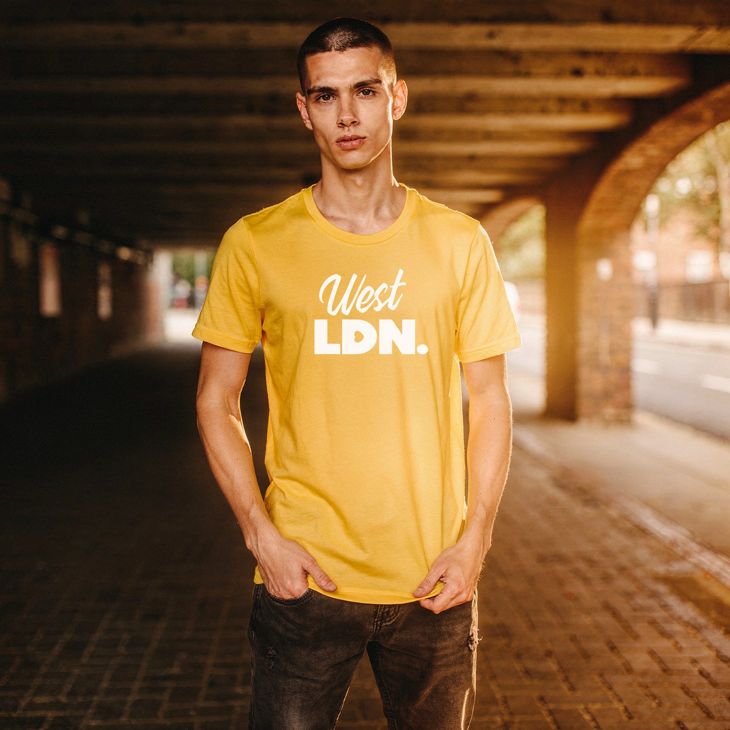 Man wearing yellow t-shirt with 'West LDN.' text, standing under a bridge golden time sun rays