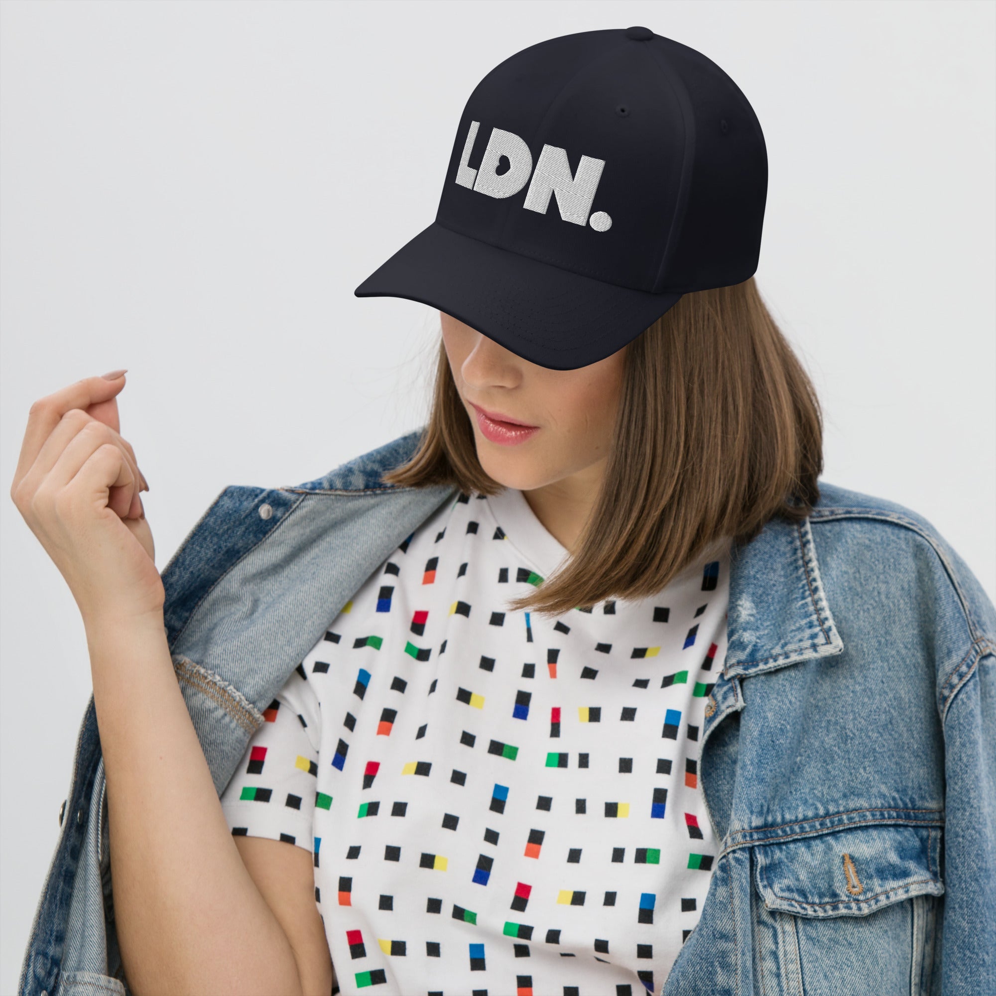 Person wearing a black cap with 'LDN.' text, white t-shirt with colorful pattern, and denim jacket on a light gray background