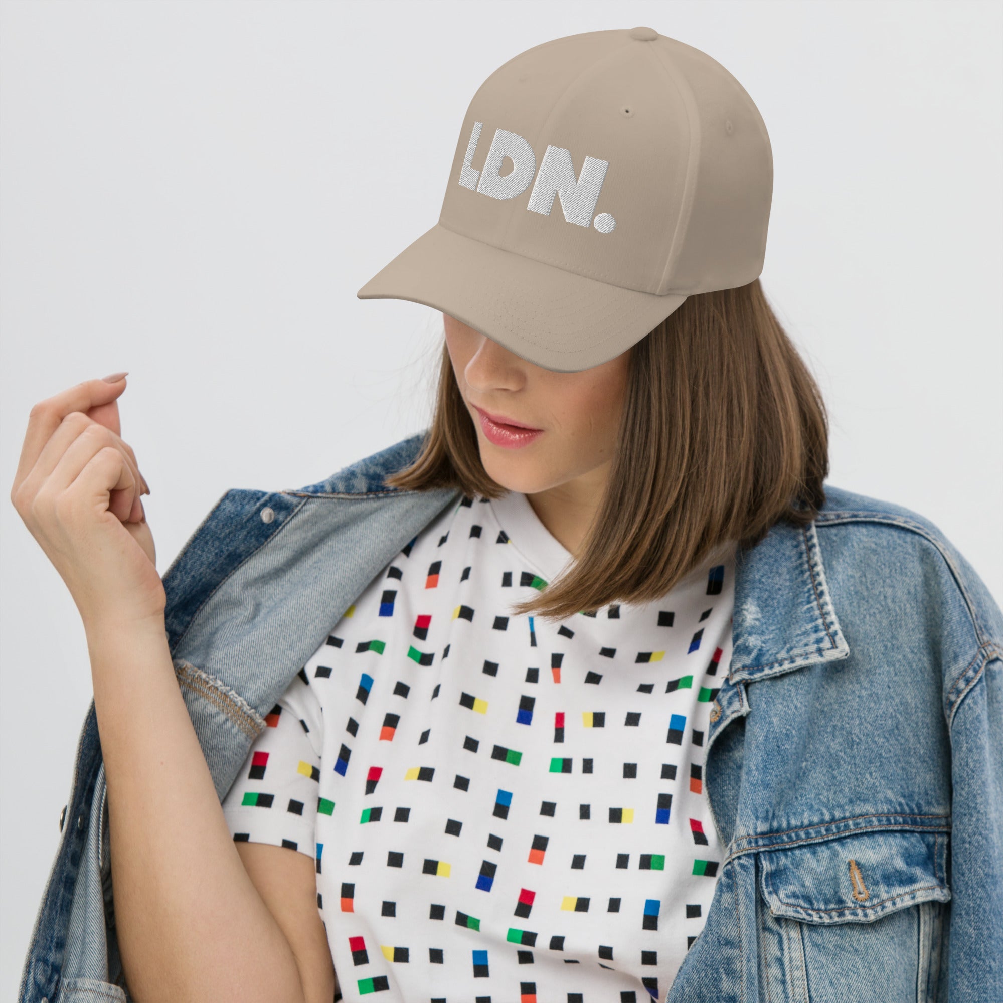 Person wearing a beige cap with 'LDN.' text, white t-shirt with colorful pattern, and denim jacket on a light gray background