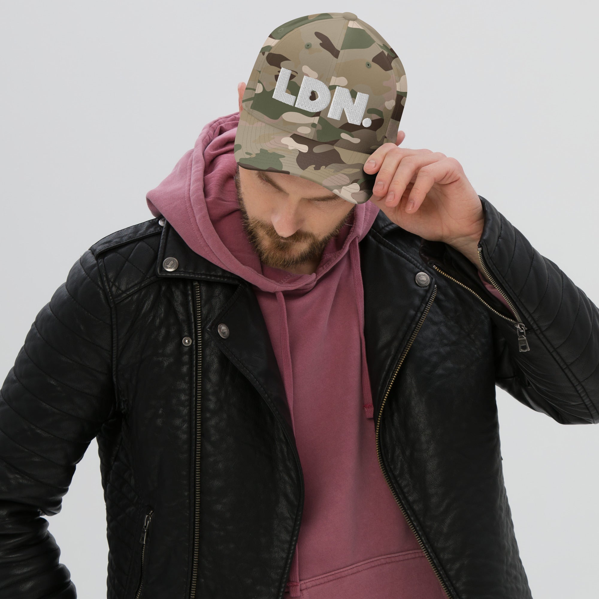 Person wearing a camouflage cap with 'LDN.' text, black leather jacket, and pink hoodie on a light gray background