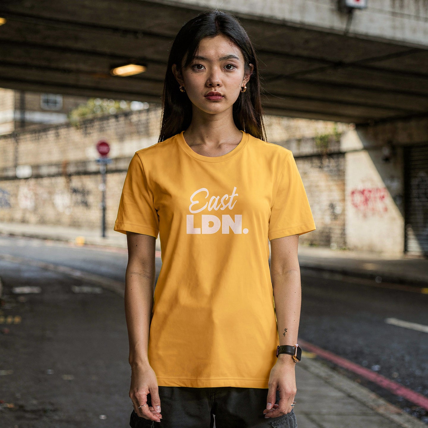 Person wearing a yellow 'East LDN.' t-shirt standing under an overpass.