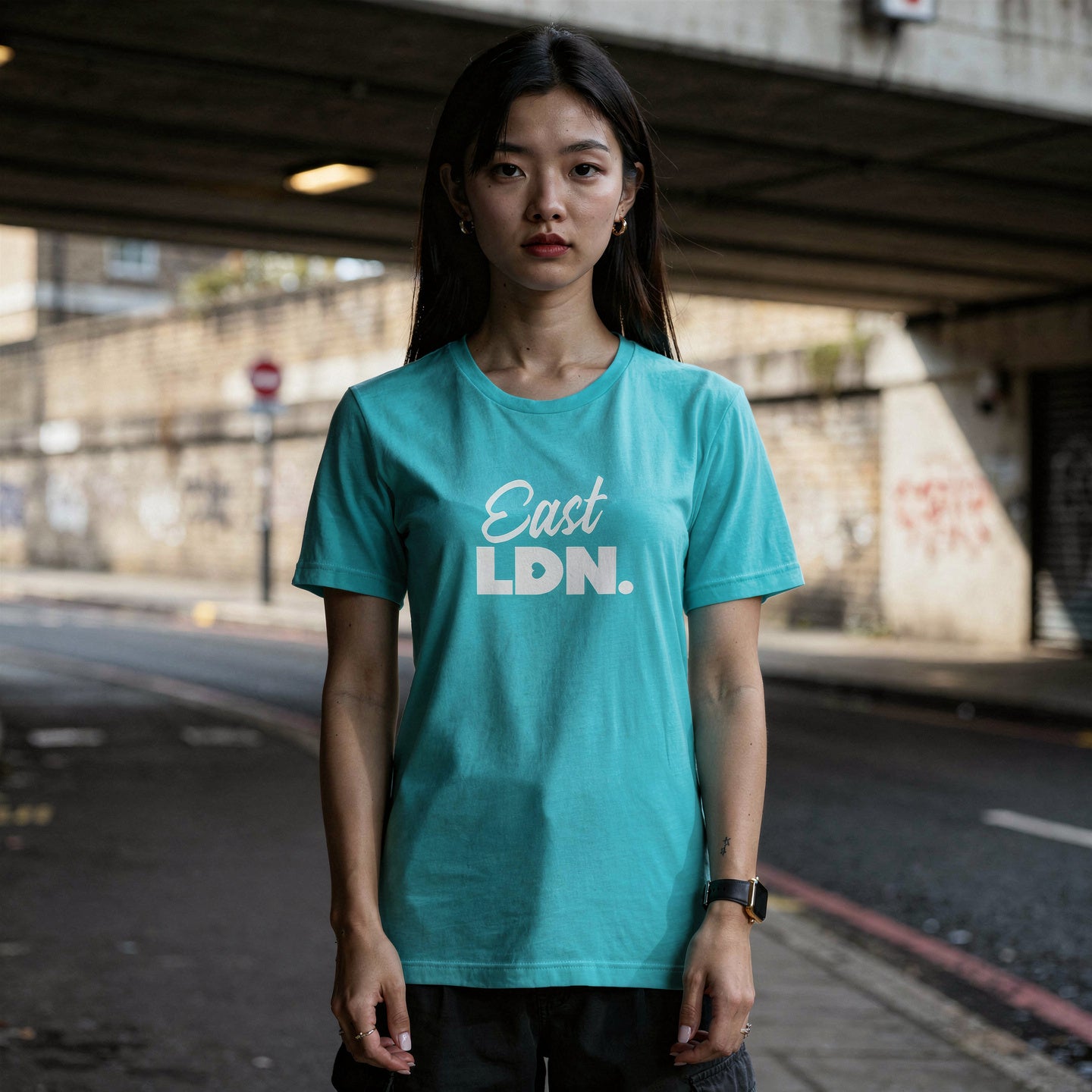 Person wearing a teal t-shirt with 'East LDN.' text standing under an overpass.