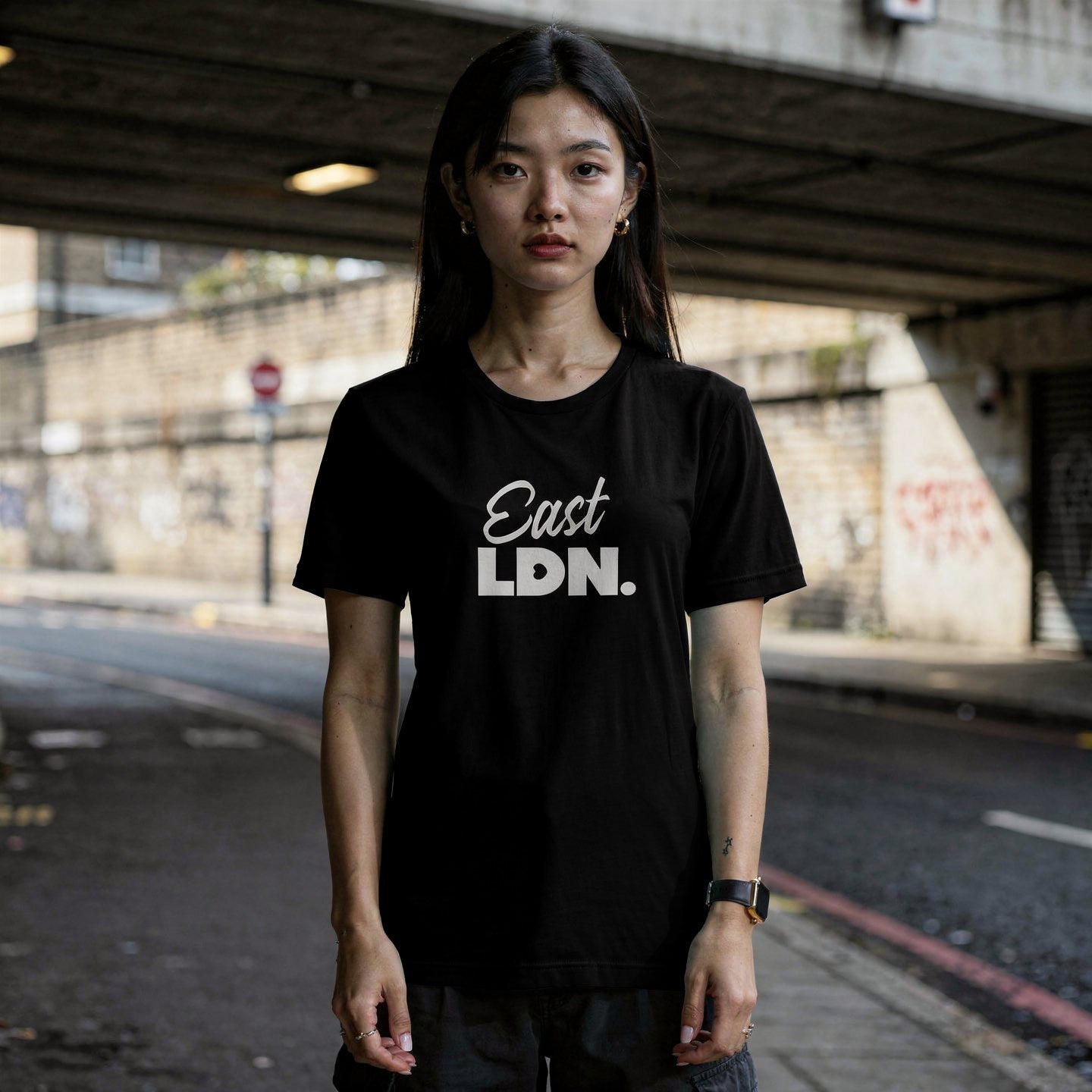 Person wearing a black t-shirt with 'East LDN' text standing under an overpass.