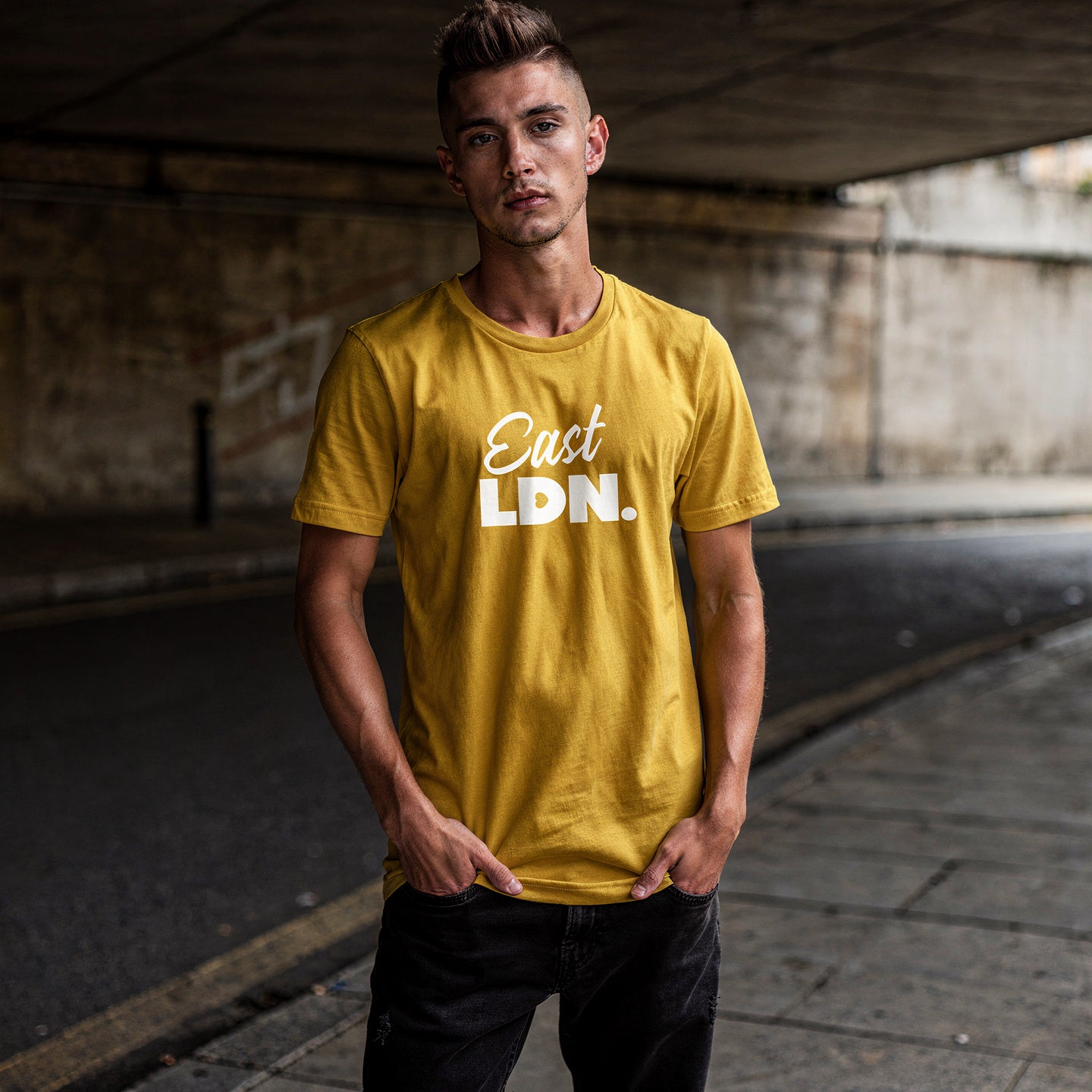 Man wearing a yellow t-shirt with 'East Ldn.' text, standing under a bridge.