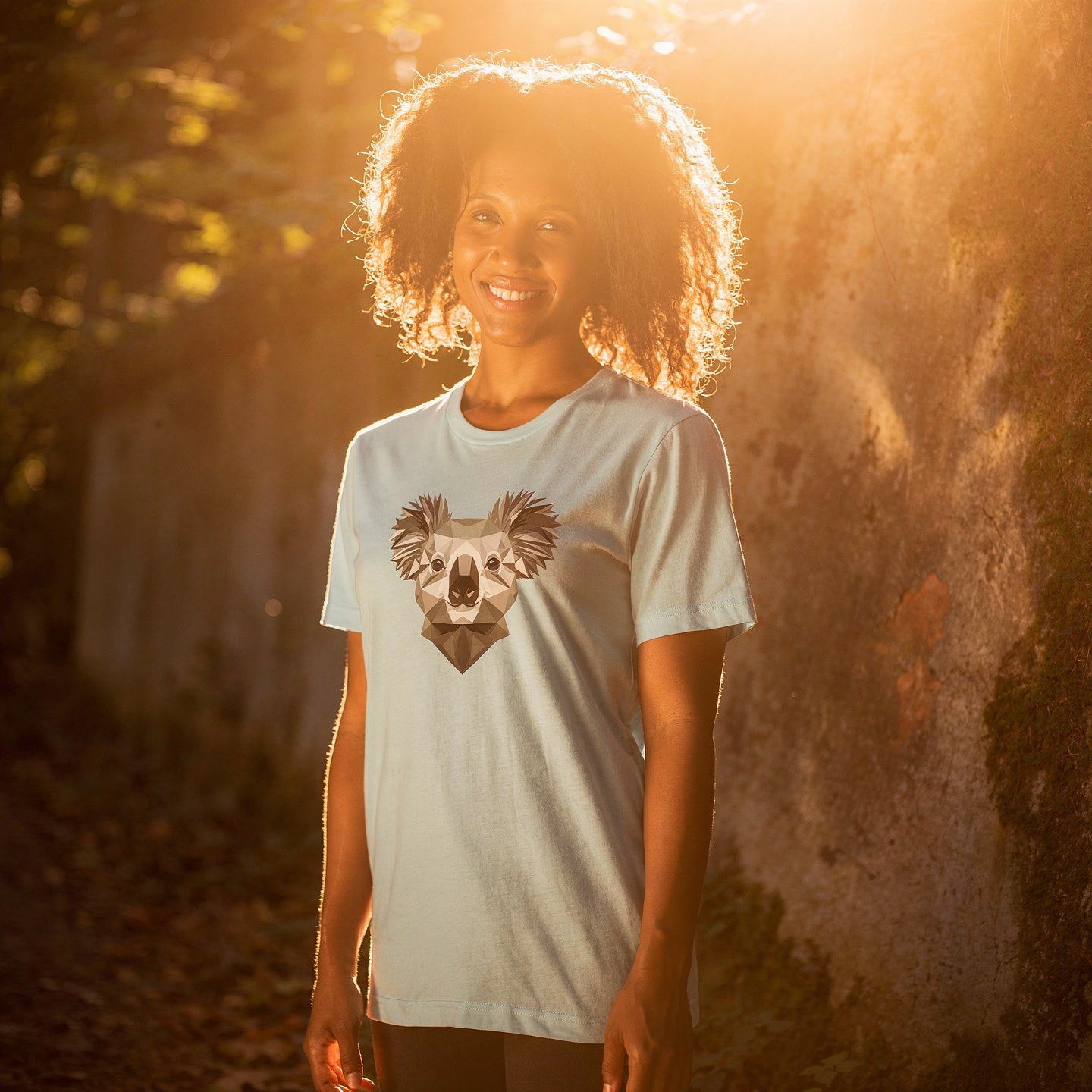 Person wearing a black t-shirt with a koala design, standing in a forest next to a large wall
