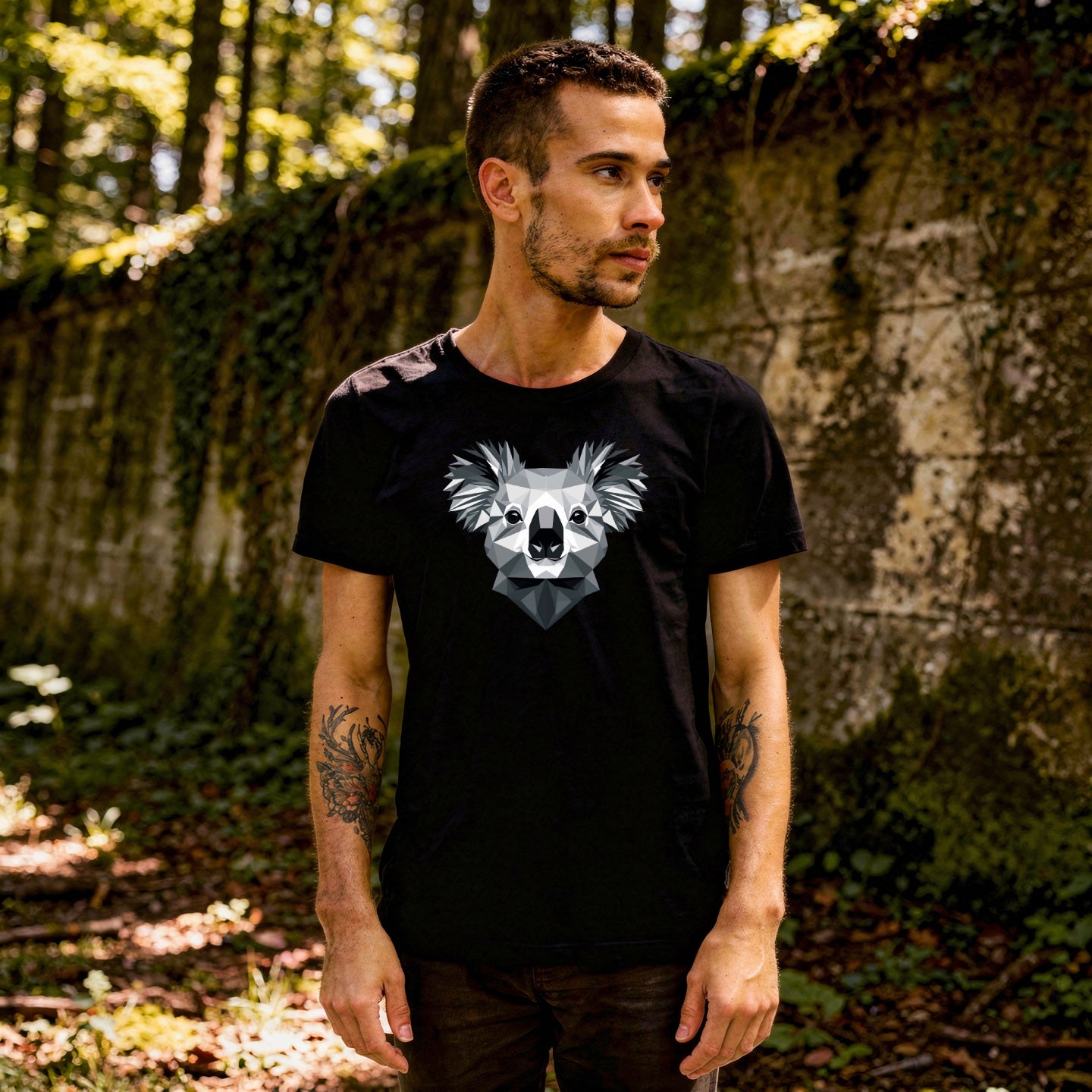 Person wearing a black t-shirt with a koala design, standing in a forest next to a large wall
