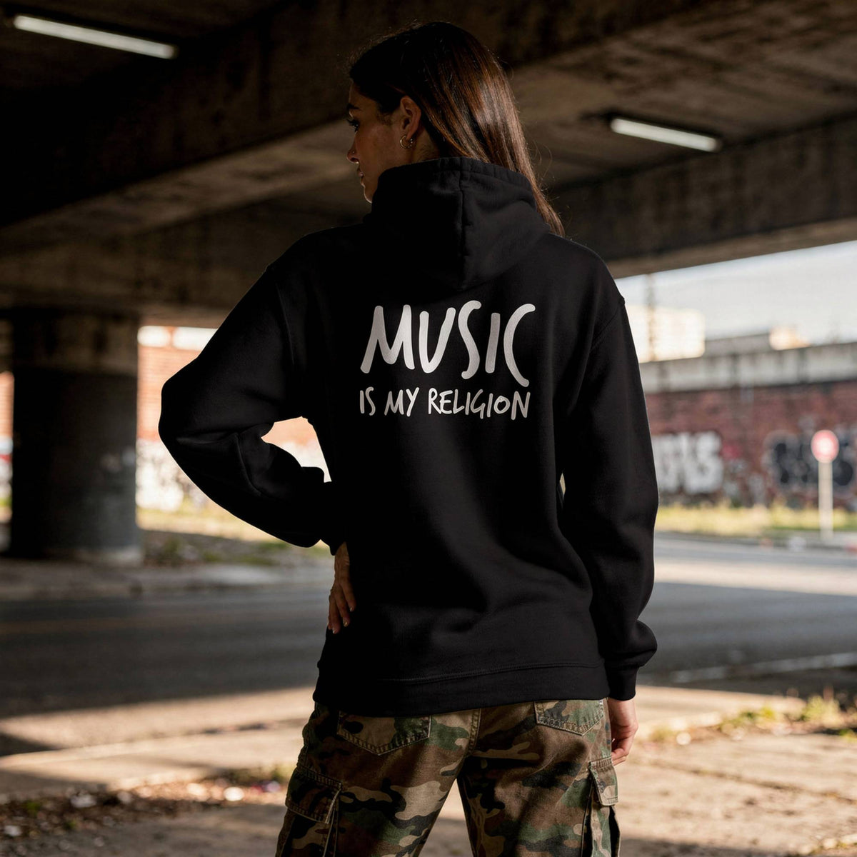 A woman stands under a concrete bridge wearing a black hoodie from the back, which features the white text 'MUSIC IS MY RELIGION' printed across the back. She is also wearing camouflage cargo pants.