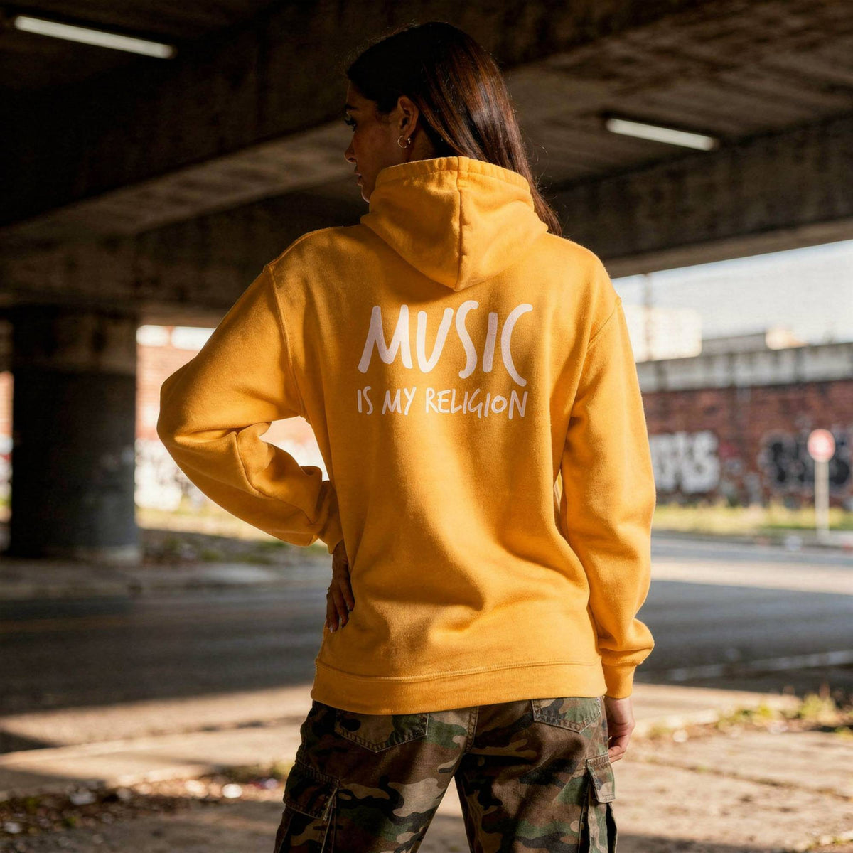 A woman stands under a concrete bridge wearing a mustard yellow hoodie with 'MUSIC IS MY RELIGION' printed in white on the back, paired with camouflage cargo pants.