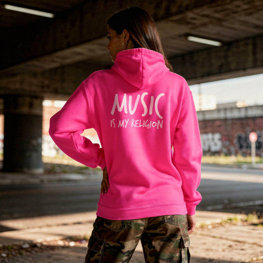 A woman stands under a concrete bridge wearing a bright pink hoodie with the phrase 'MUSIC IS MY RELIGION' printed in white across the back, paired with camouflage cargo pants.