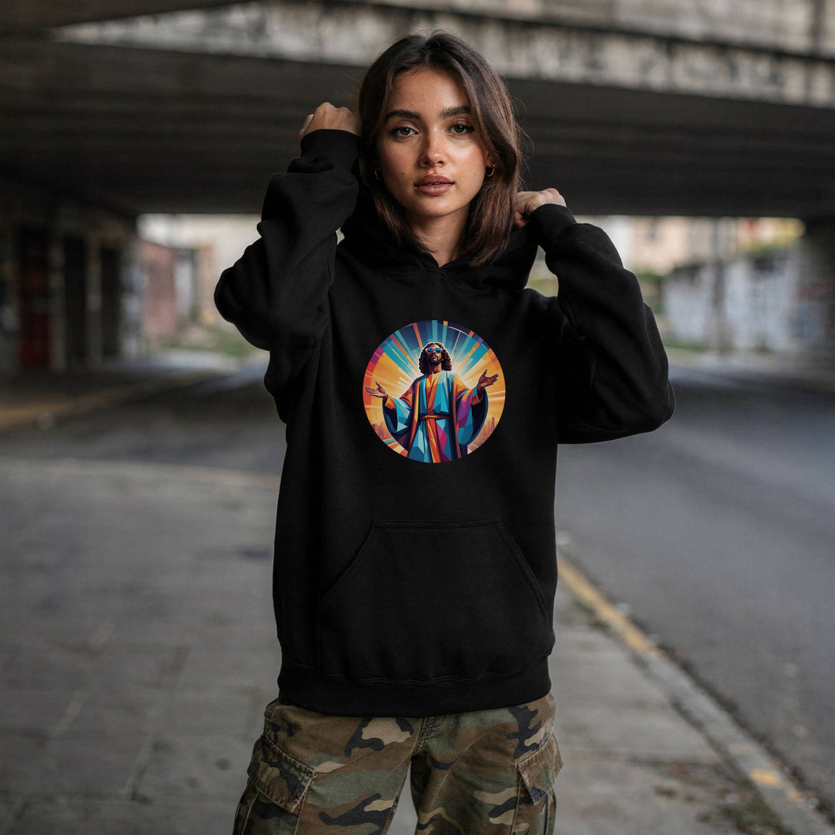 A woman wearing a black hoodie with a colorful circular graphic of Jesus on the chest, standing in an urban underpass environment.