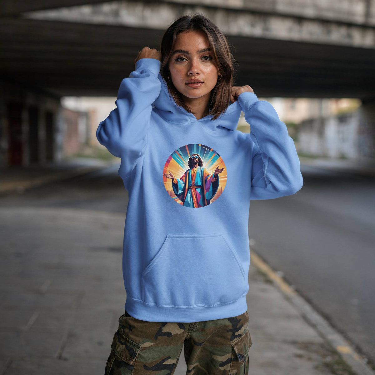 A woman wearing a light blue hoodie with a colorful circular graphic of Jesus on the chest, paired with camouflage cargo pants, standing under a concrete bridge.