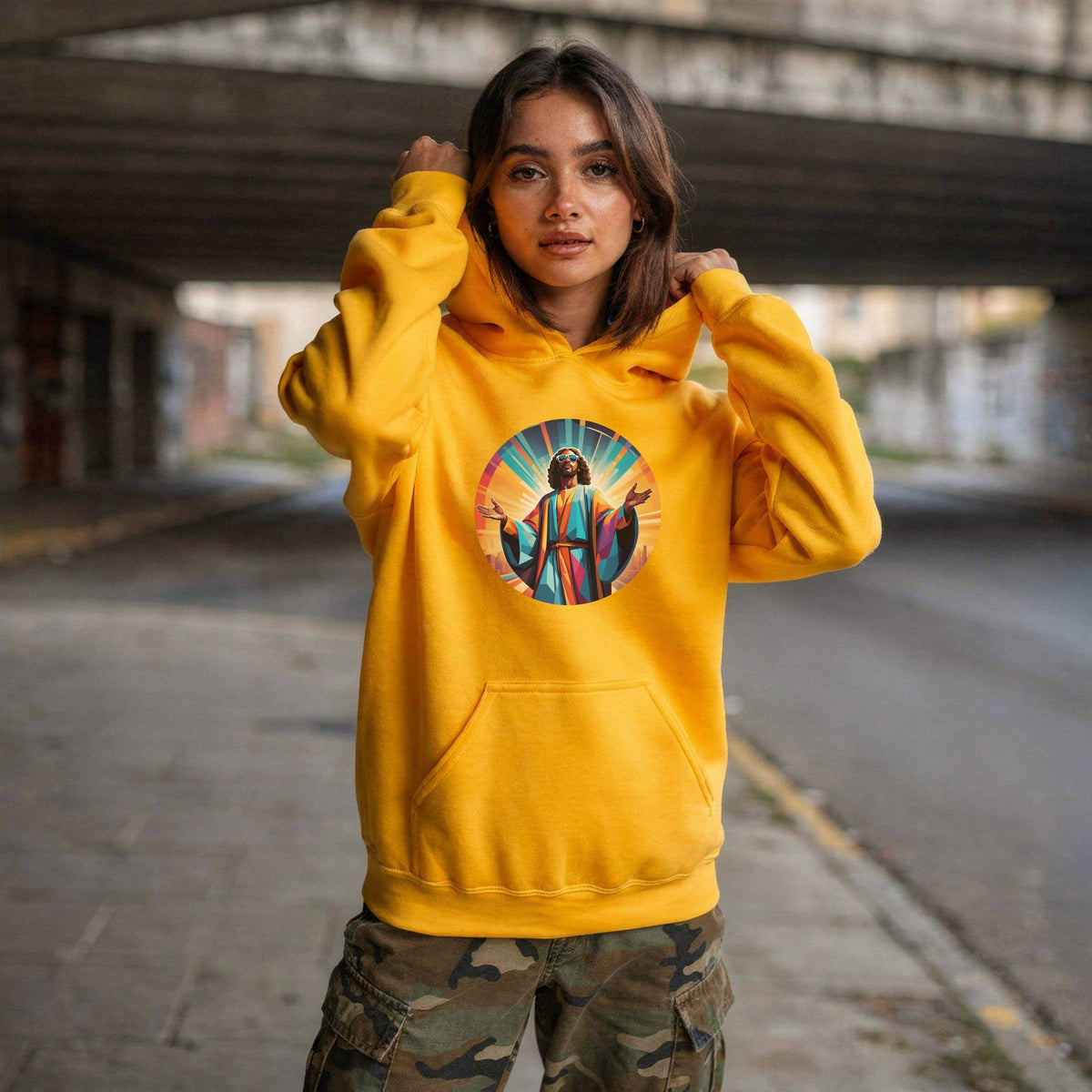 A woman wearing a bright yellow hoodie with a colorful circular graphic of Jesus on the chest, paired with camouflage cargo pants, standing in an urban alleyway under a concrete bridge.