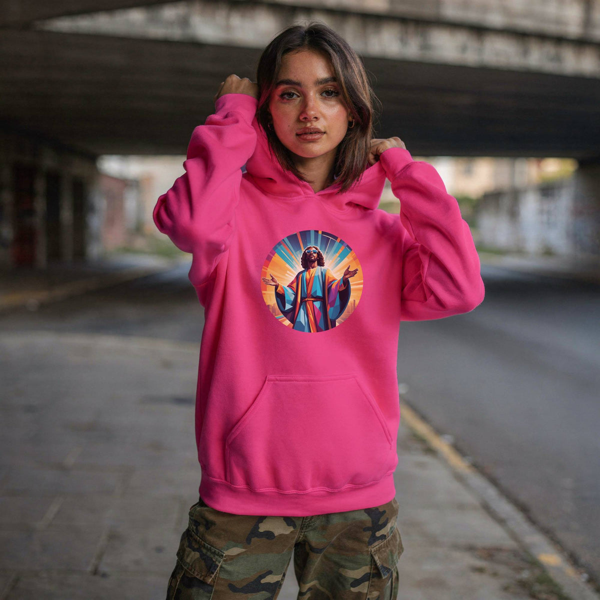 A woman wearing a bright pink hoodie with a colorful circular graphic of Jesus on the chest, posing on a street under a concrete bridge.