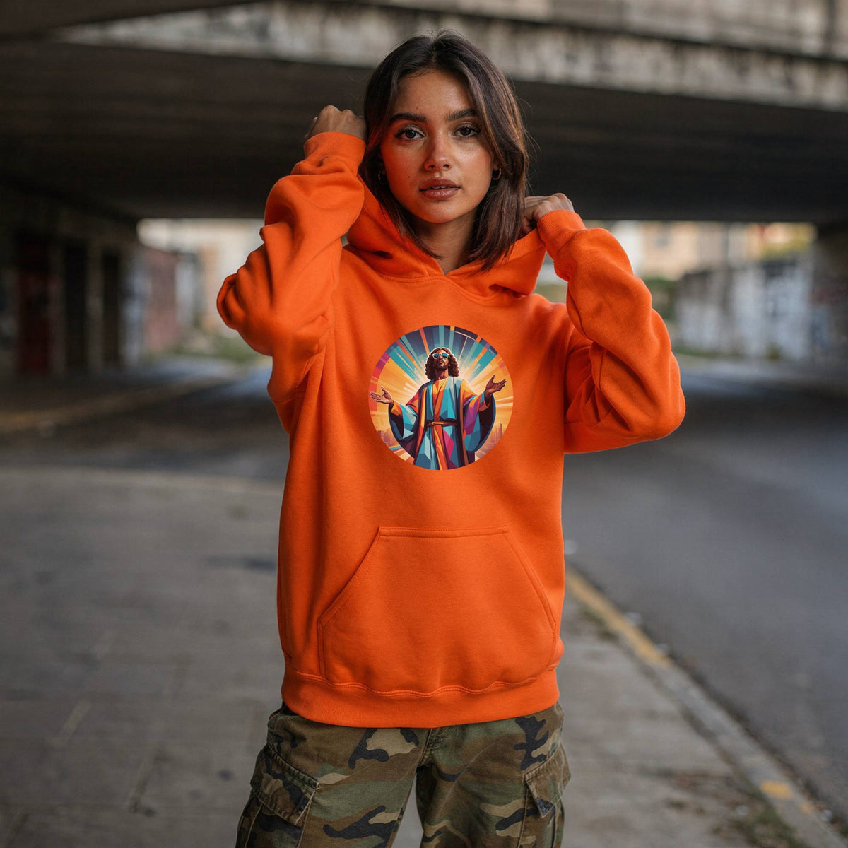 A woman wearing a bright orange hoodie with a colorful circular graphic of Jesus on the chest, standing in an urban setting under a concrete bridge.