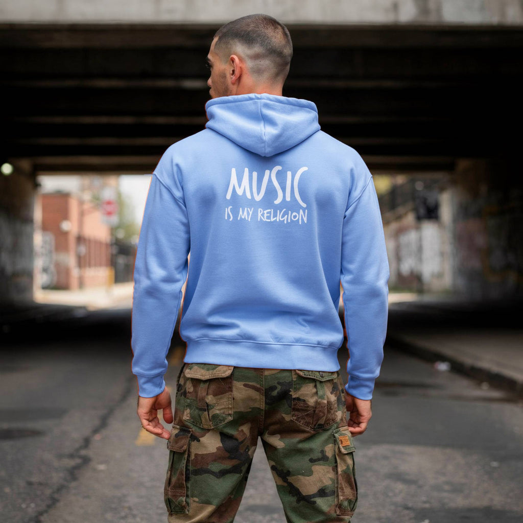 A man viewed from behind wearing a light blue hoodie with the text 'MUSIC IS MY RELIGION' printed in white on the back, paired with camouflage cargo pants, standing in an urban underpass.