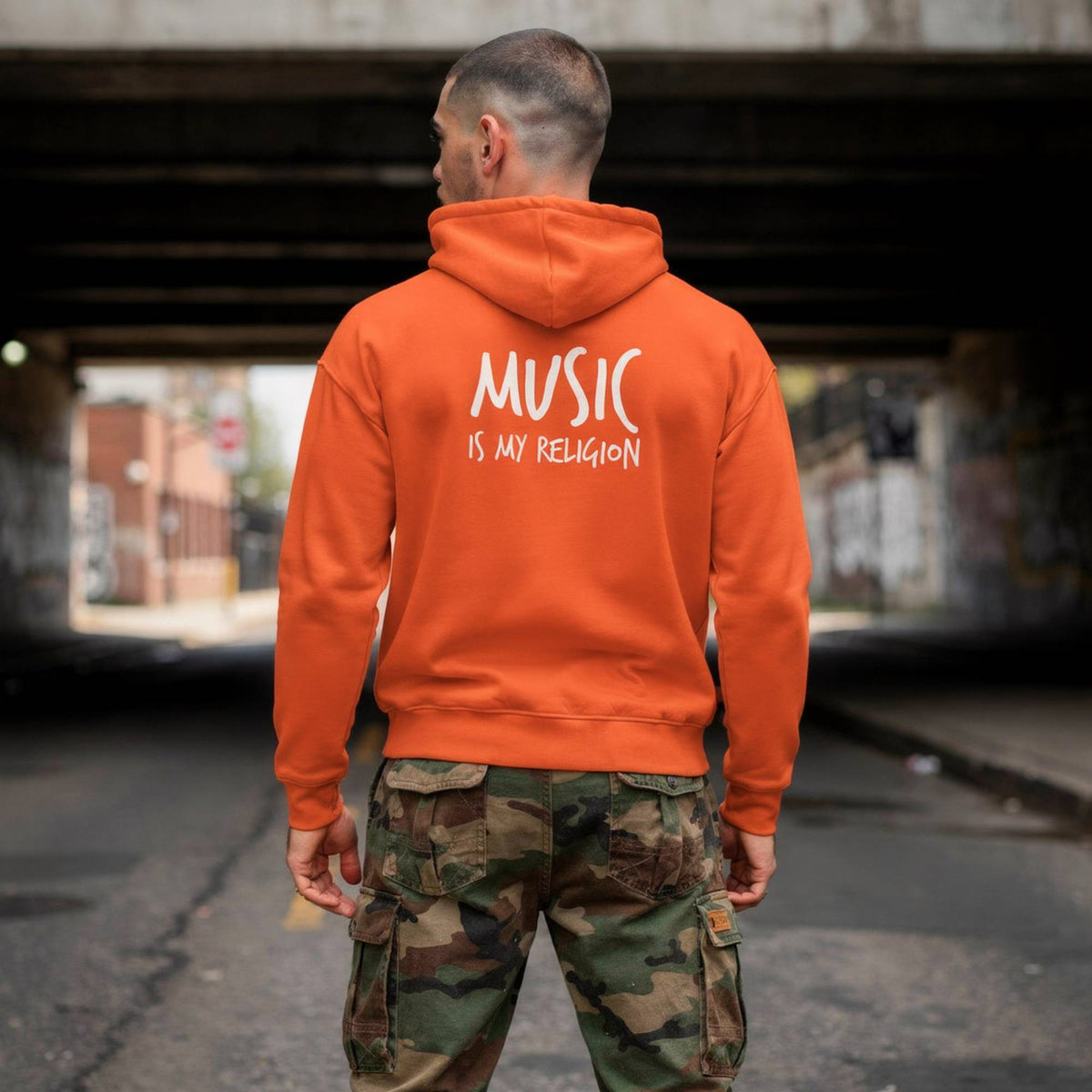 A man viewed from behind wearing a bright orange hoodie with the white text 'MUSIC IS MY RELIGION' printed on the back, paired with camouflage cargo pants, standing under a concrete bridge.