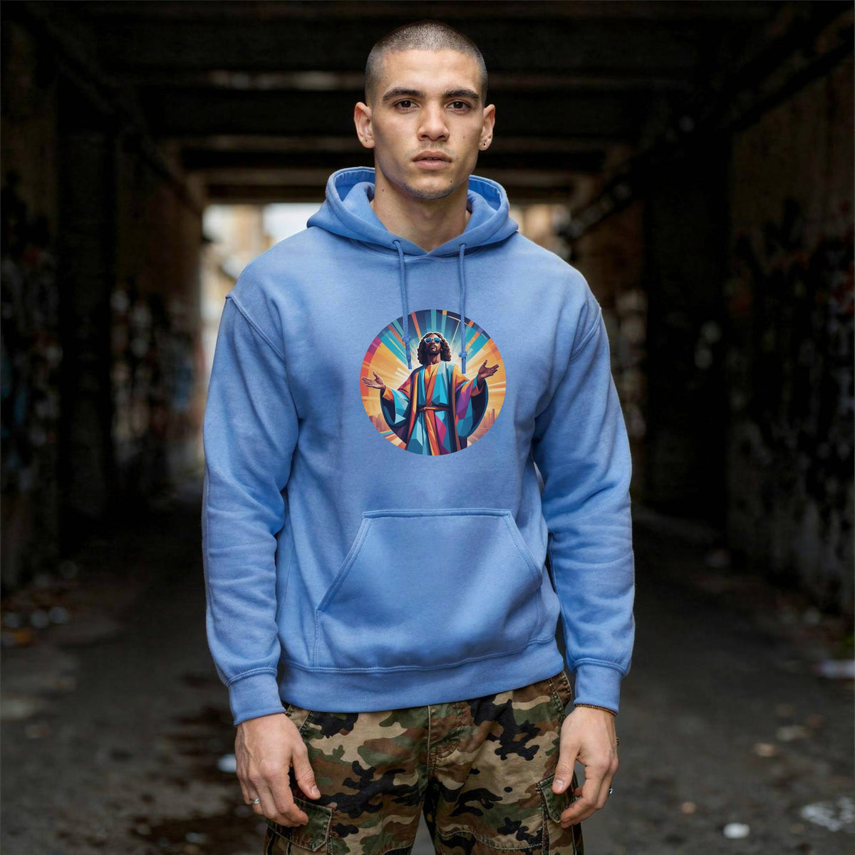 A man wearing a light blue hoodie with a colorful graphic of Jesus Christ on the chest, standing in a dark, graffiti-covered alleyway.