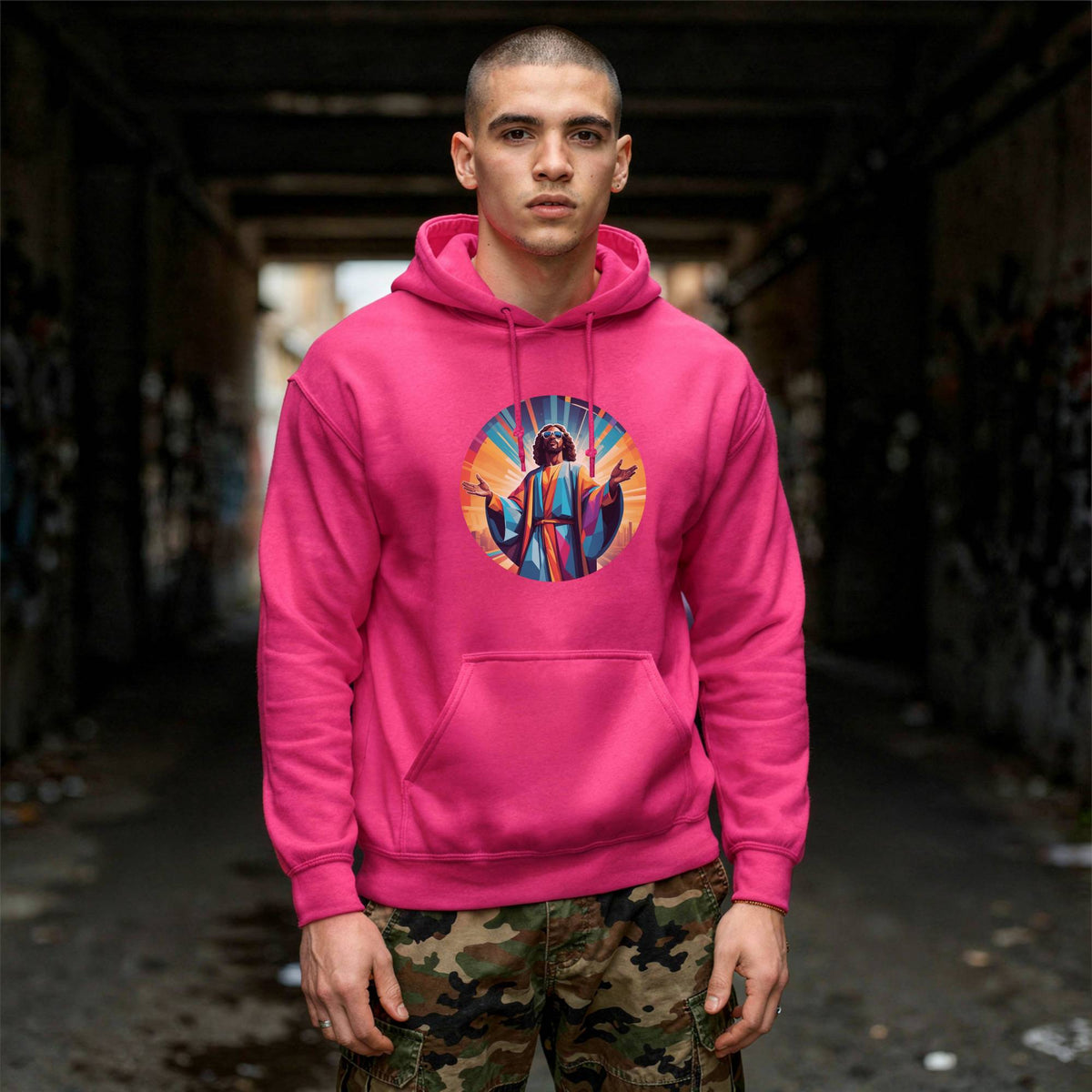 A man wearing a bright pink pullover hoodie with a colorful circular graphic of Funky Jesus Christ on the chest, standing in a dark, graffiti-covered alleyway.