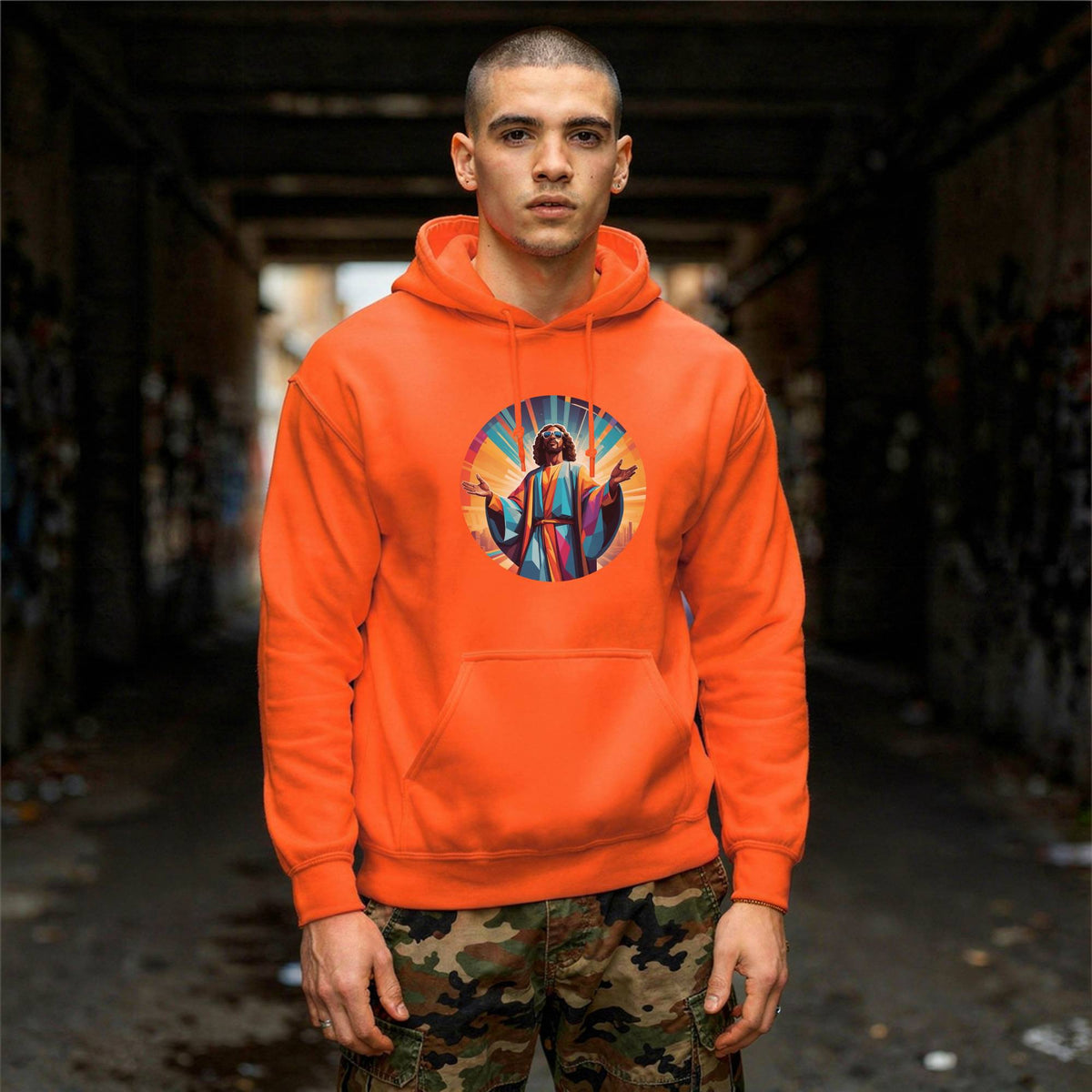 A man wearing a bright orange hoodie with a colorful circular graphic of Jesus on the chest, standing in a dark, graffiti-covered alleyway.