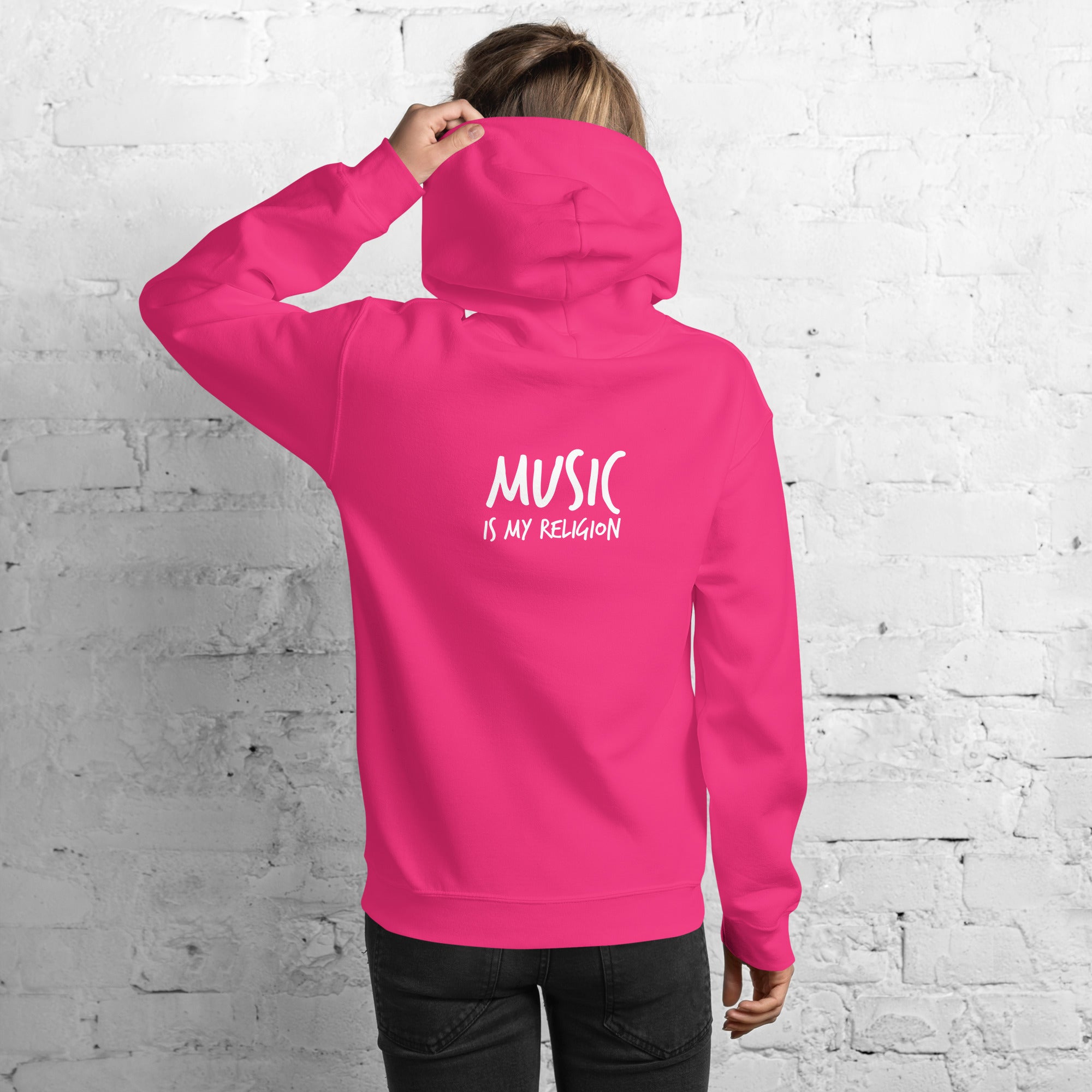 Person wearing a pink hoodie with 'Music is my Religion' text on the back against a white brick wall.