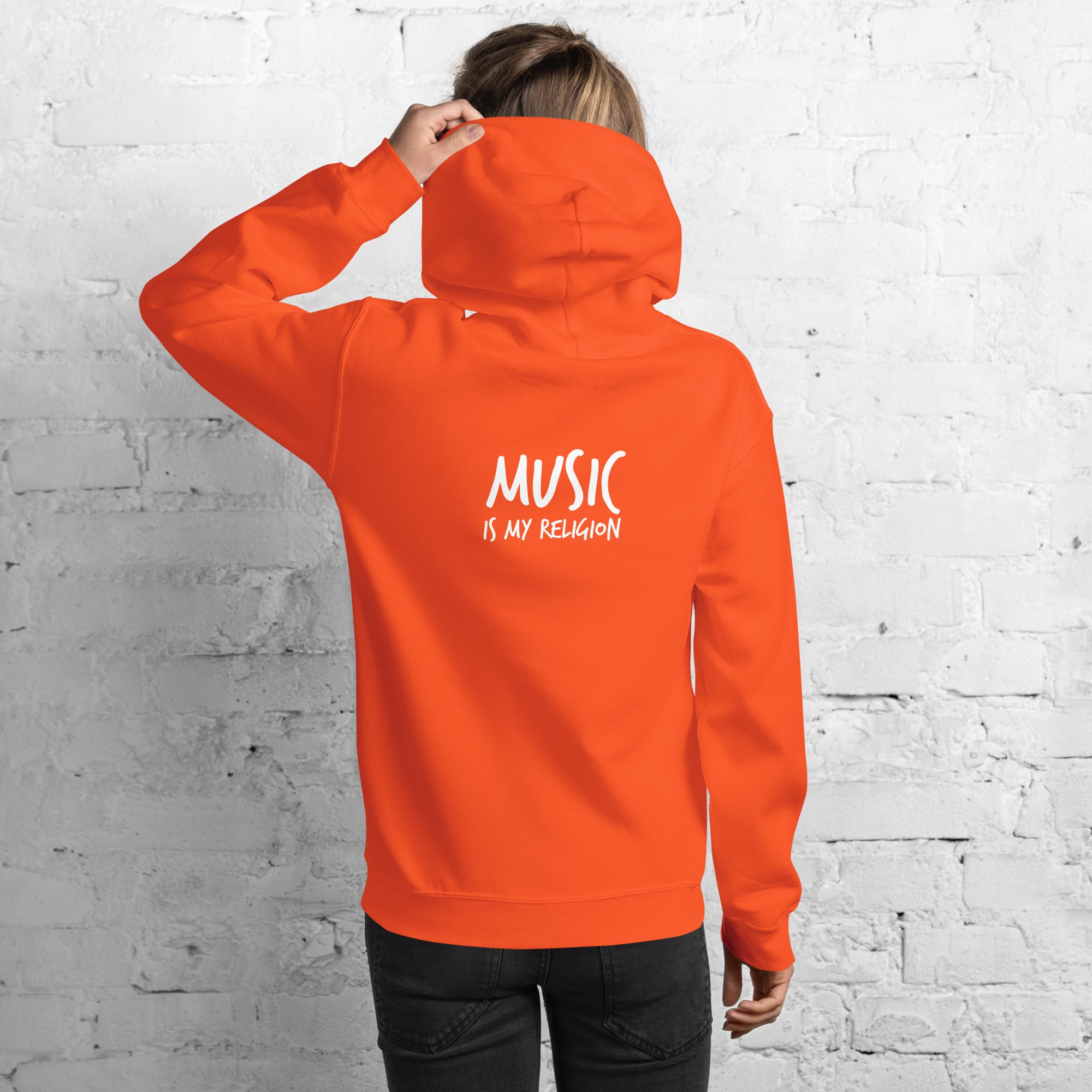 Person wearing an orange hoodie with 'Music is my Religion' text on the back against a white brick wall.