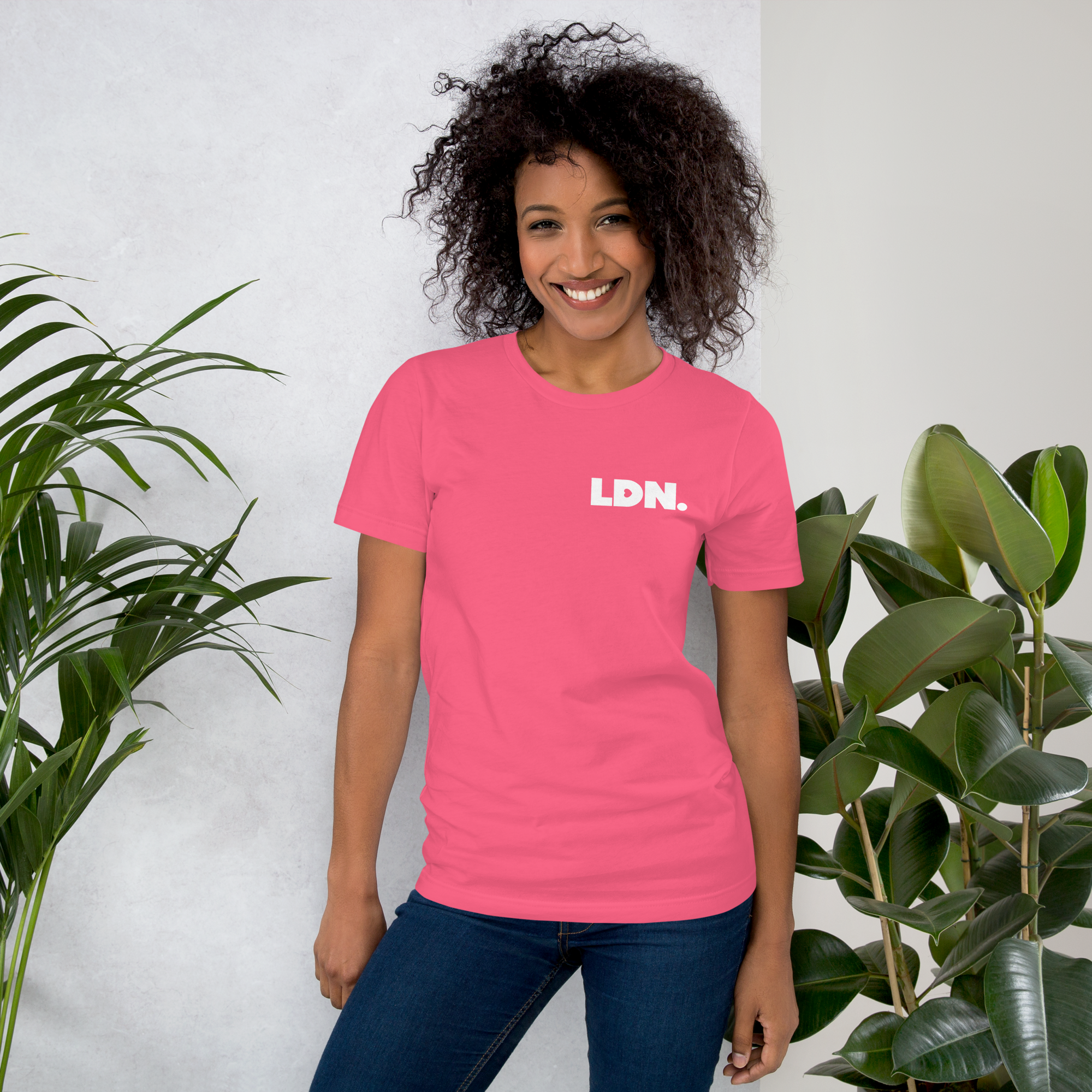 Woman wearing a pink t-shirt with 'LDN.' text, standing between two plants against a white wall.