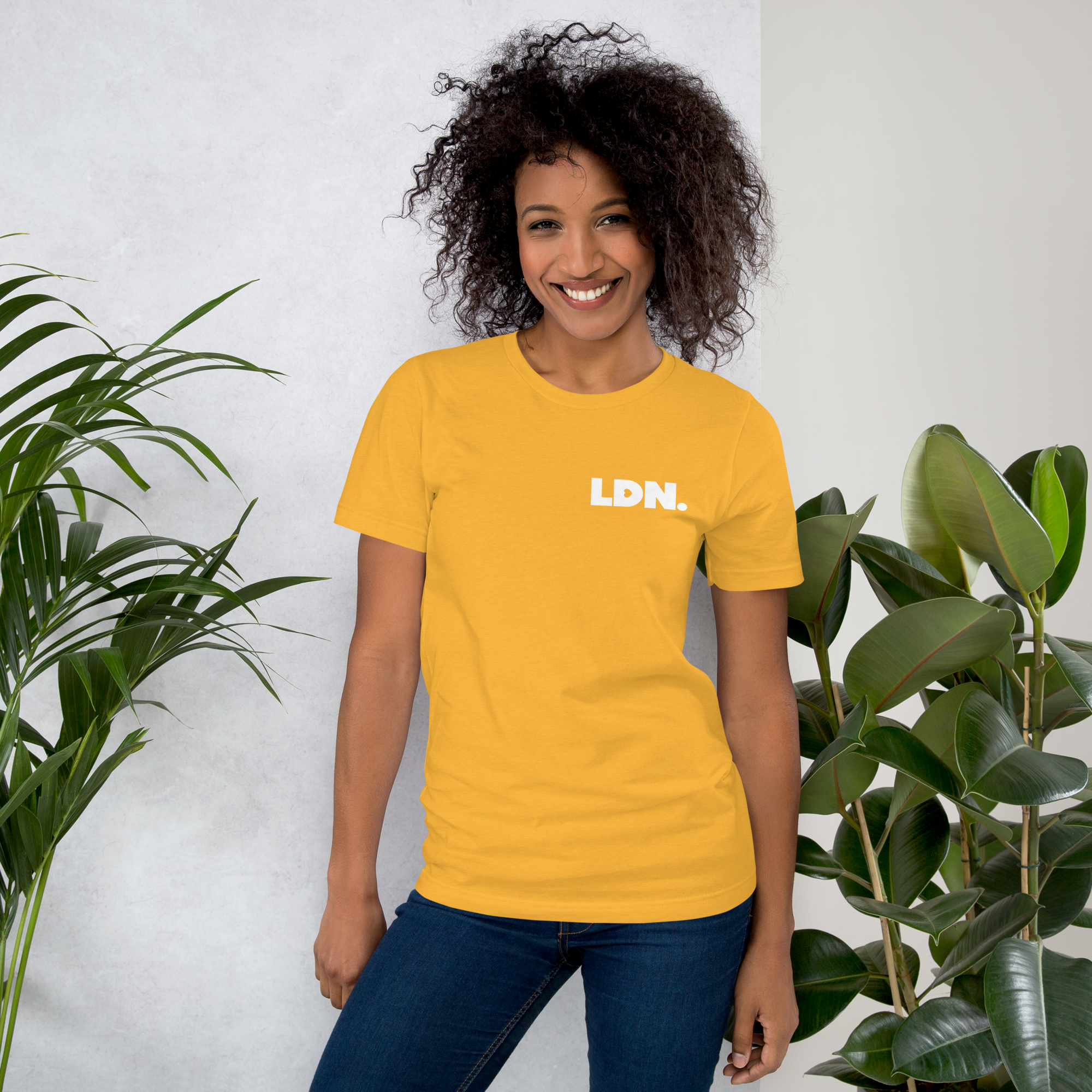 Person wearing a yellow t-shirt with 'LDN.' text, standing between two plants against a white wall.