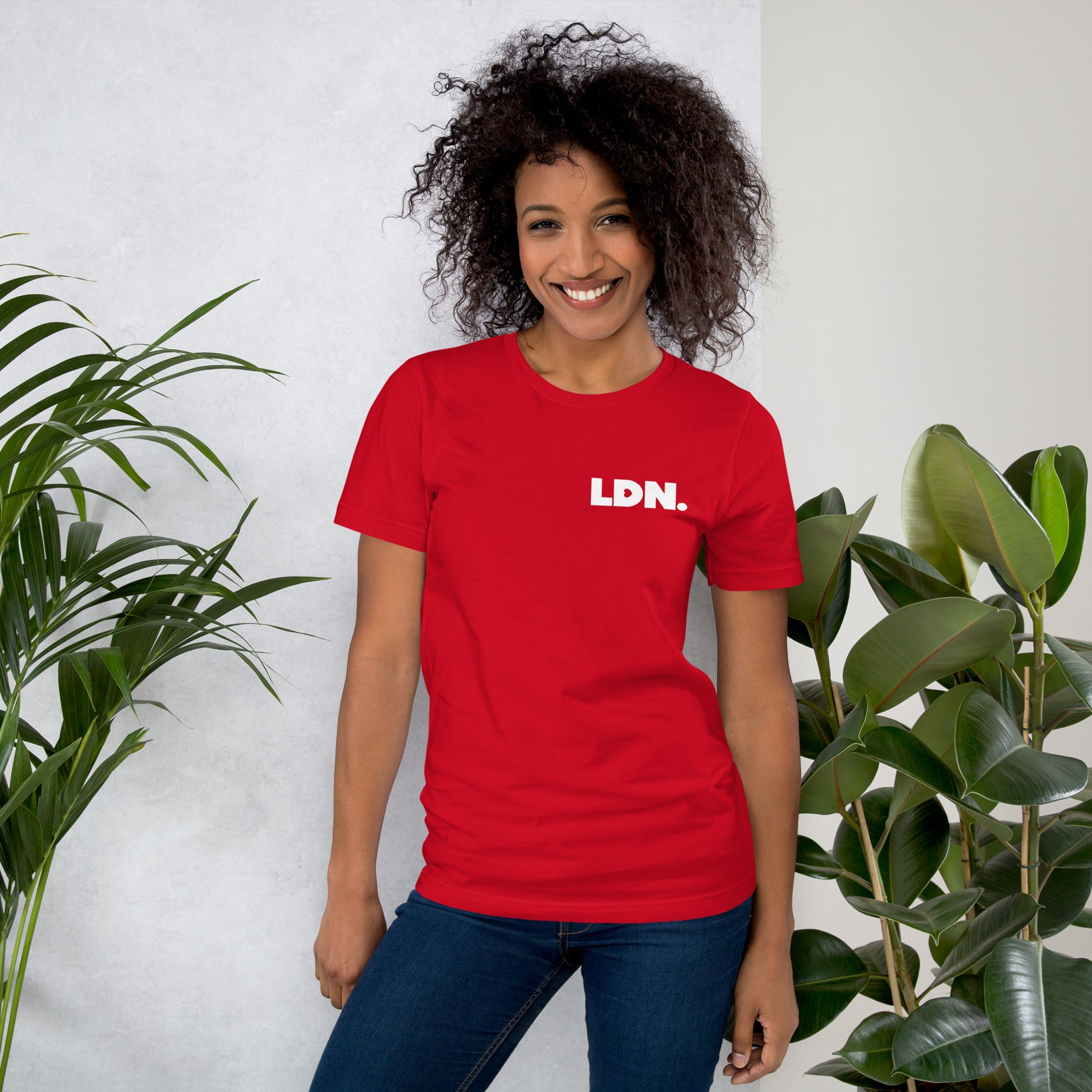 Person wearing a red t-shirt with 'LDN' text, standing between two plants against a white wall.