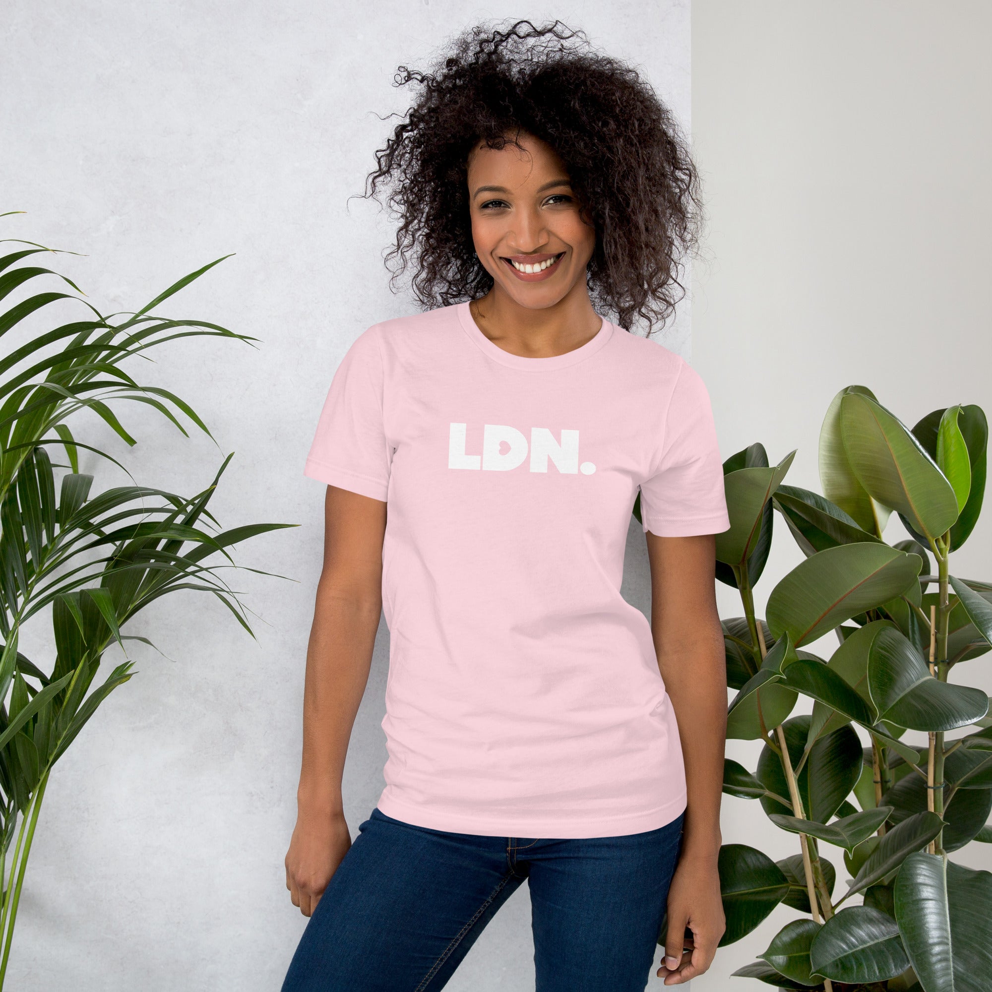 Person wearing a pink t-shirt with 'LDN.' text, standing between two plants against a light background