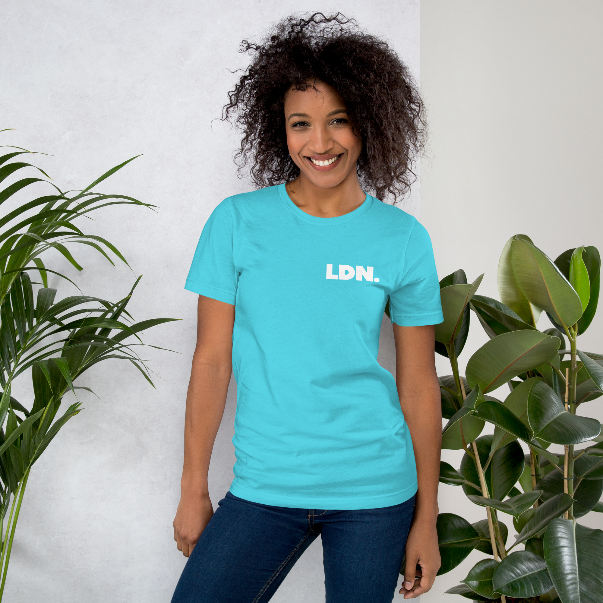 Woman wearing a turquoise t-shirt with 'LDN.' text, standing between two plants against a white wall.