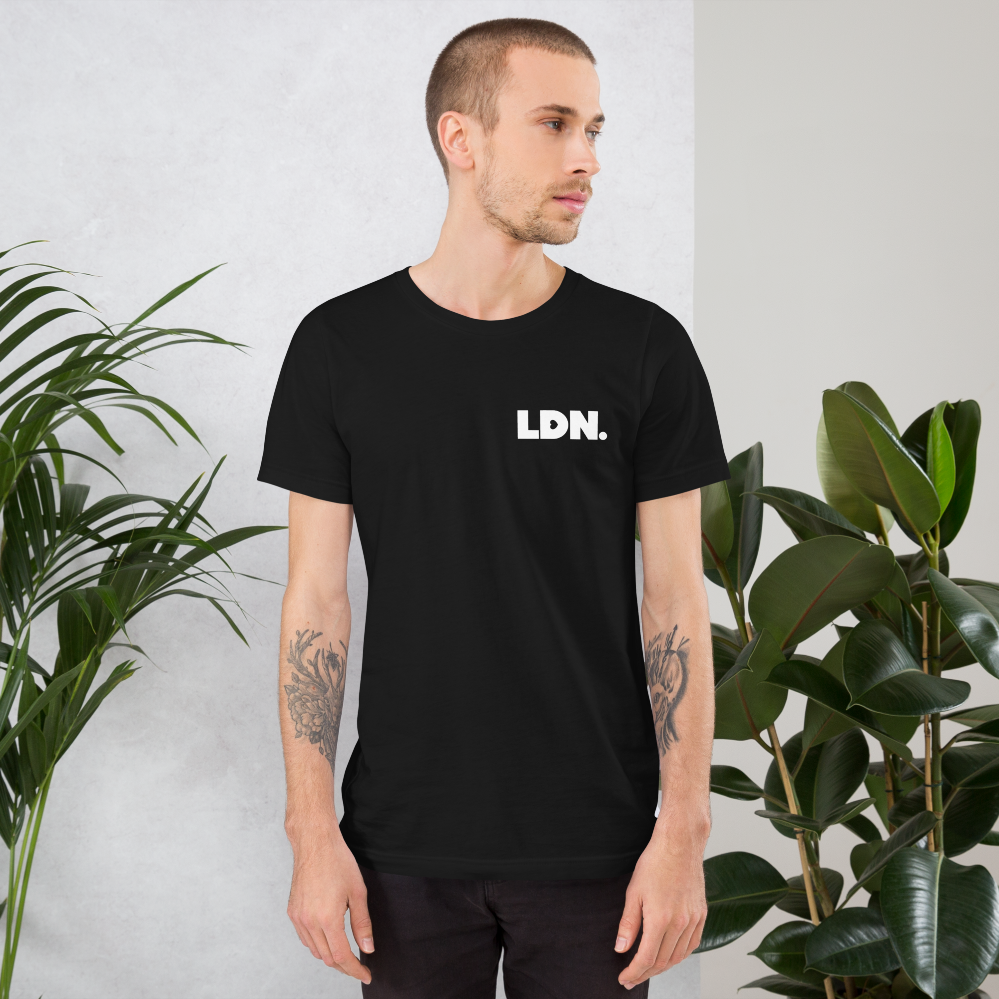 Man wearing a black t-shirt with 'LDN.' text, standing between two potted plants.