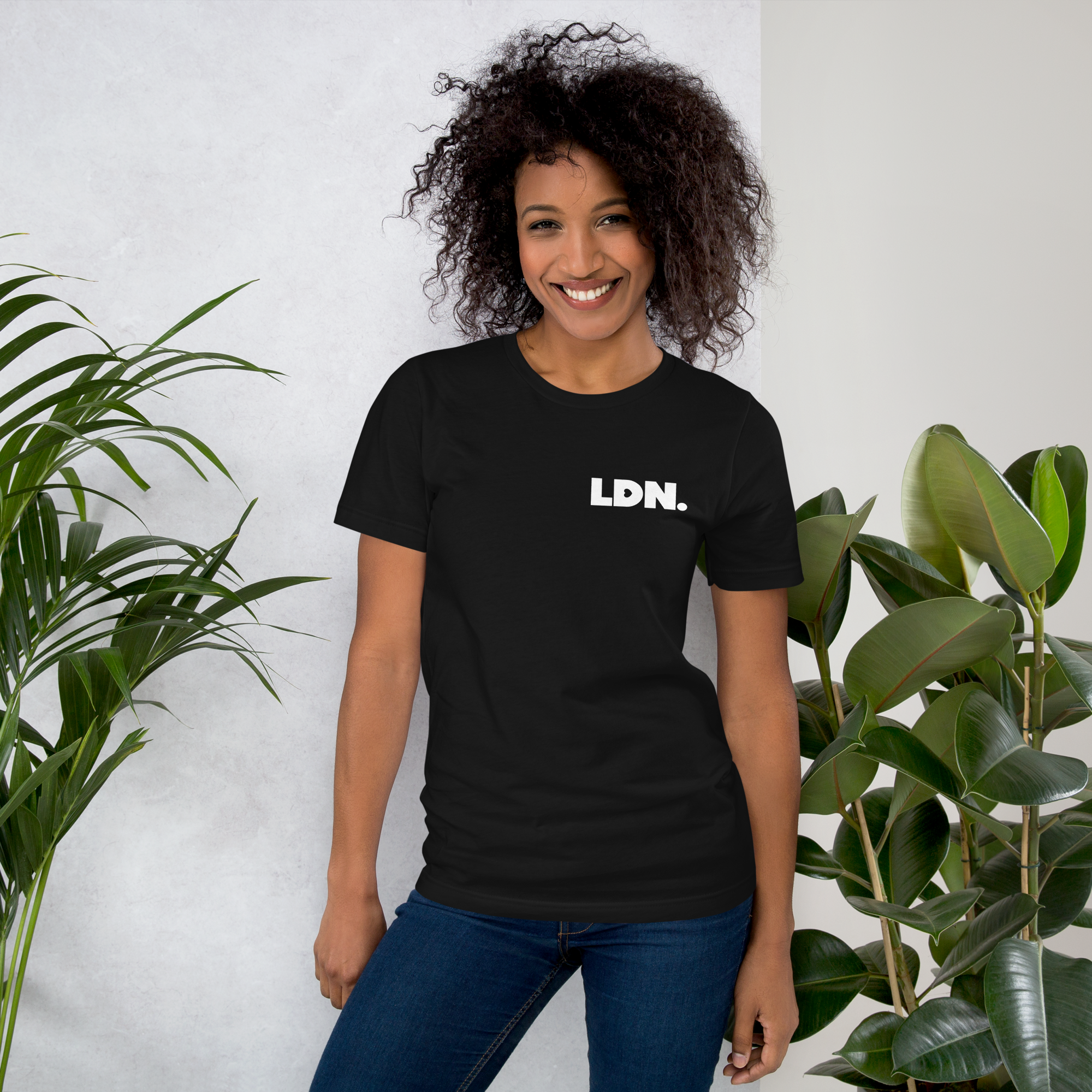 Person wearing a black t-shirt with 'LDN.' text, standing between two plants against a white wall.
