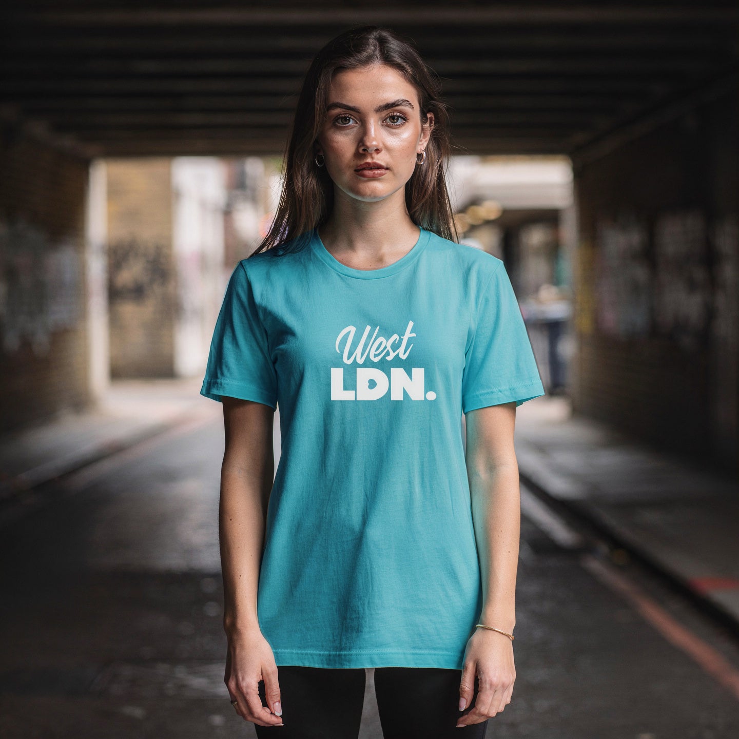 woman wearing a turquoise t-shirt with 'West LDN.' text, standing under a bridge with graffiti.