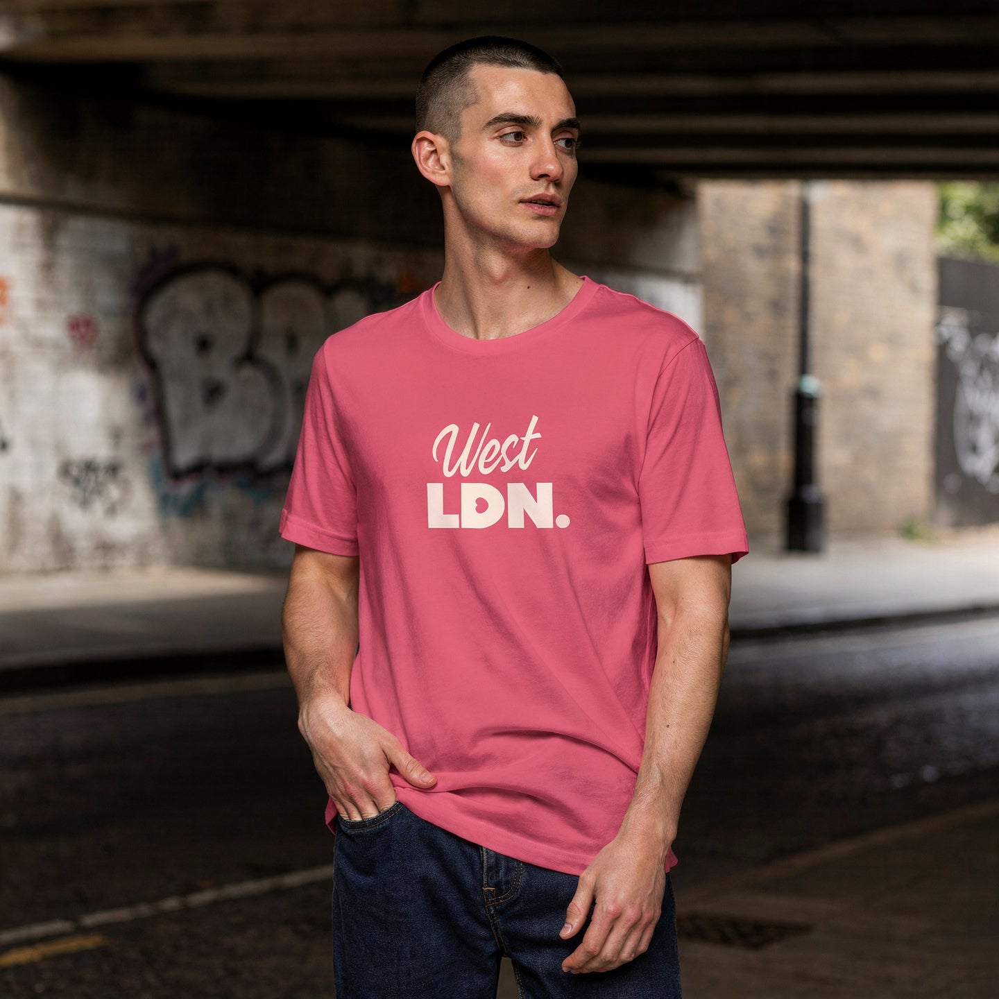 Man wearing a pink t-shirt with 'West LDN.' text, standing under an overpass with graffiti.