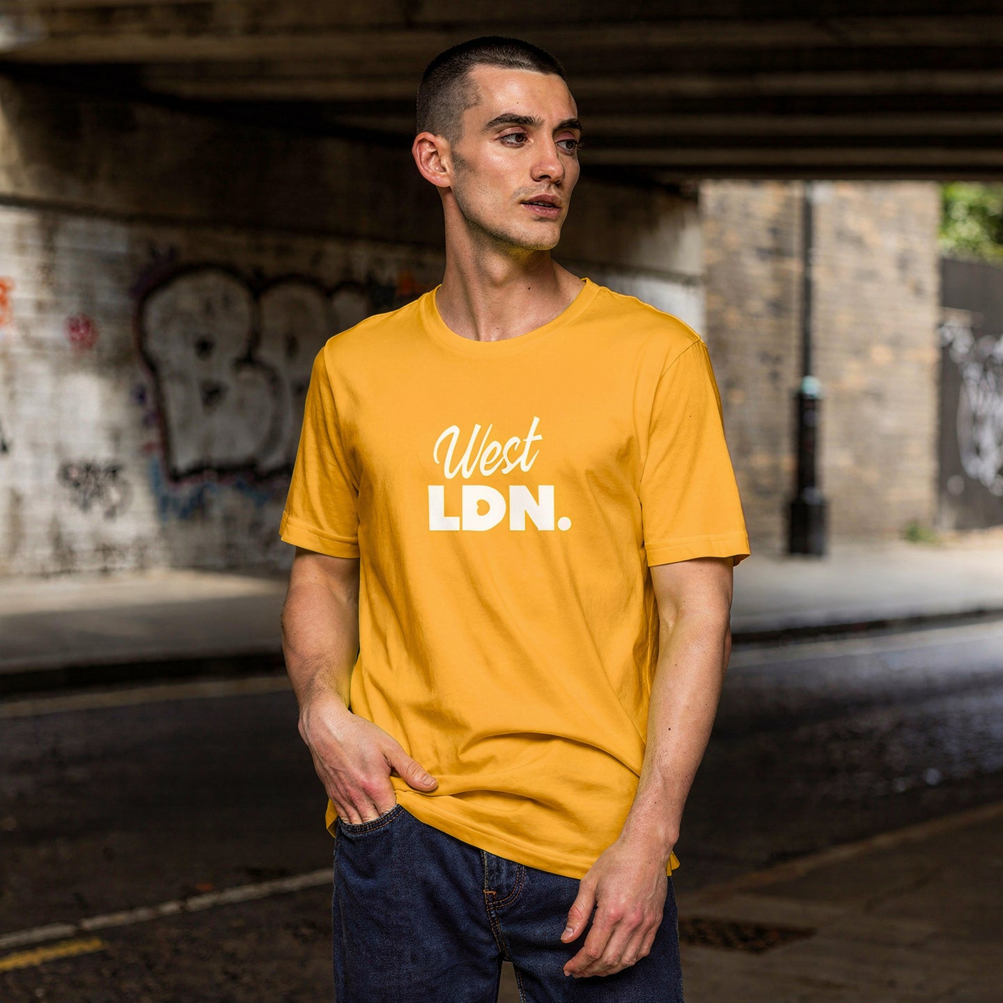 Man wearing a yellow t-shirt with 'West LDN' text standing under a bridge with graffiti.