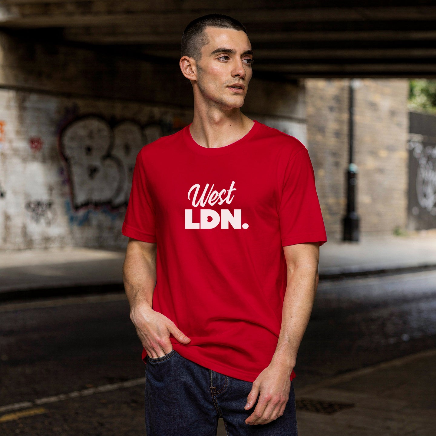 Man wearing a red t-shirt with 'West LDN.' text, standing under a bridge with graffiti.