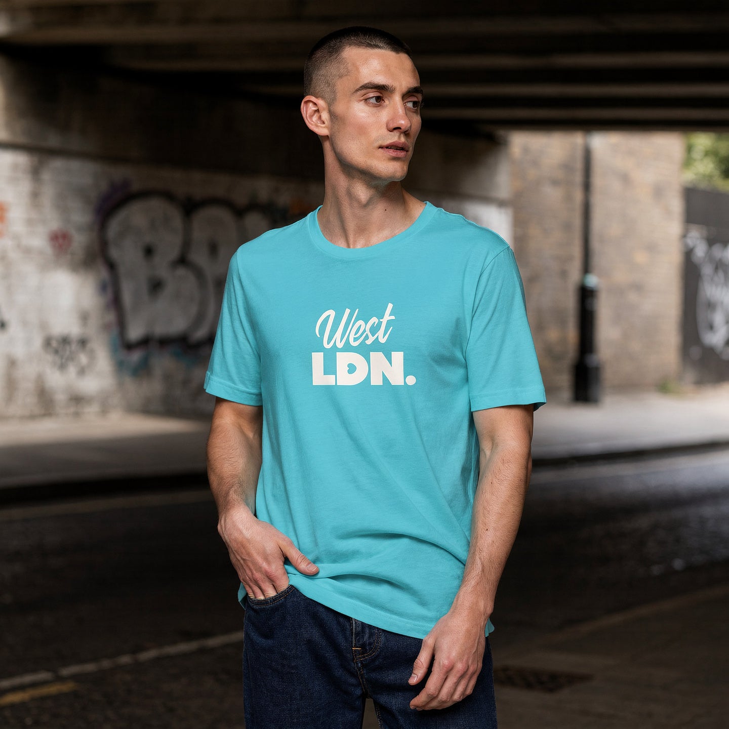 Man wearing a turquoise t-shirt with 'West LDN.' text, standing under a bridge with graffiti.