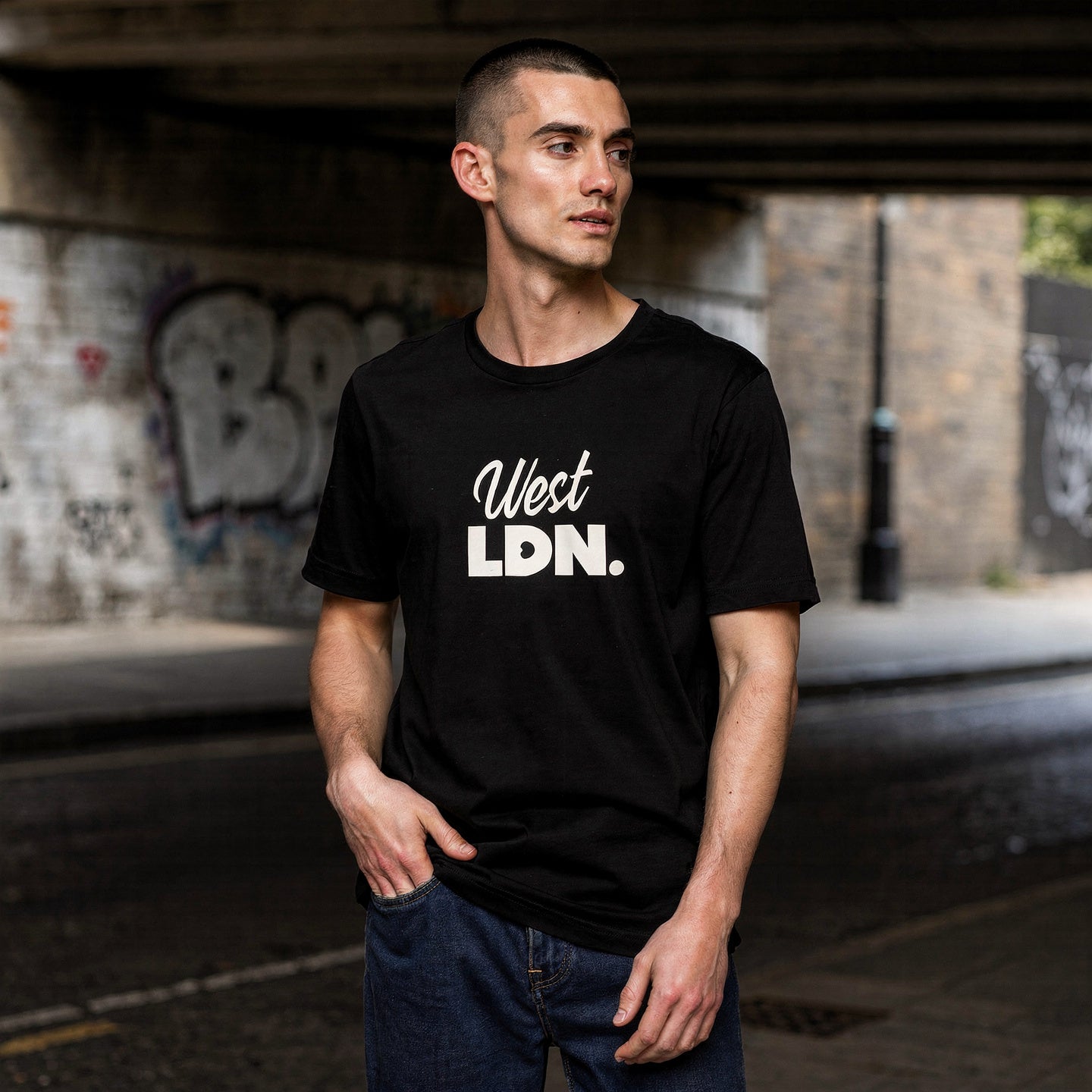 Man wearing a black t-shirt with 'West LDN.' text standing under a bridge with graffiti.