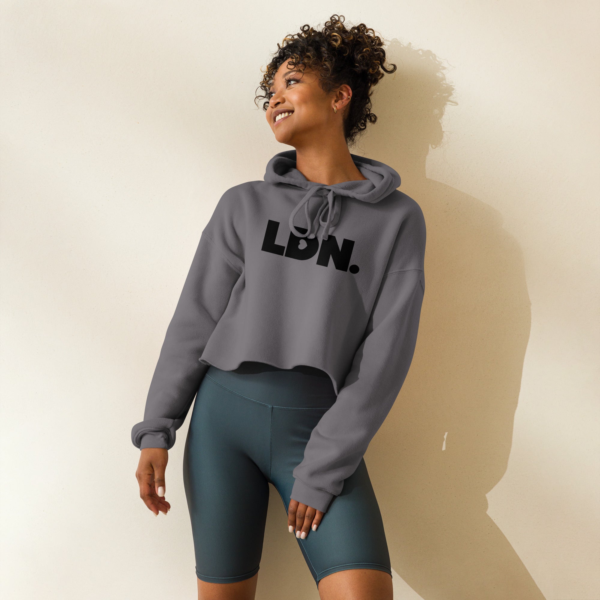 Model wearing a cropped hoodie with tonal “LDN.” graphic. Hoodie features dropped shoulders, raw hem, and dyed-to-match drawstrings. Made from cotton-poly fleece blend for soft comfort and relaxed fit. A minimal, modern streetwear piece.